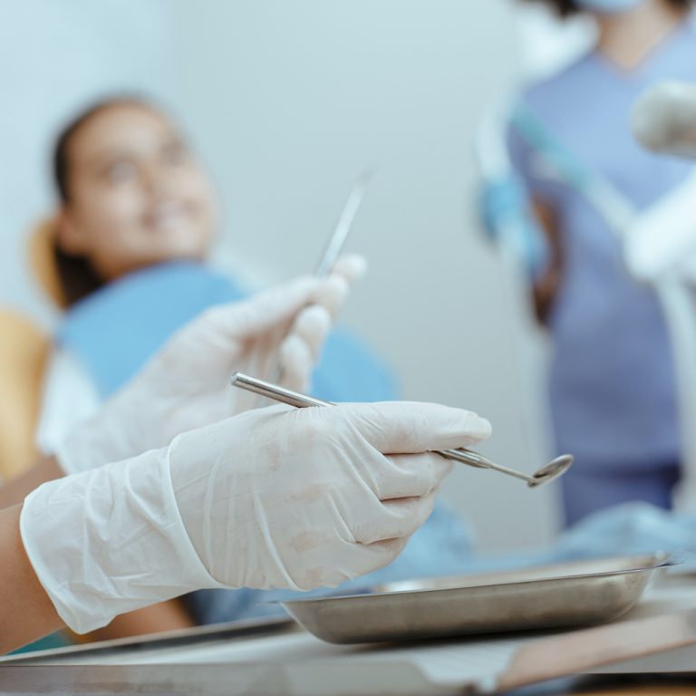 Treatment of toothache in children in modern dental clinic. Focus on hands of dentist in rubber gloves holds medical instruments with hispanic girl and nurse in blurred background, cropped, free space