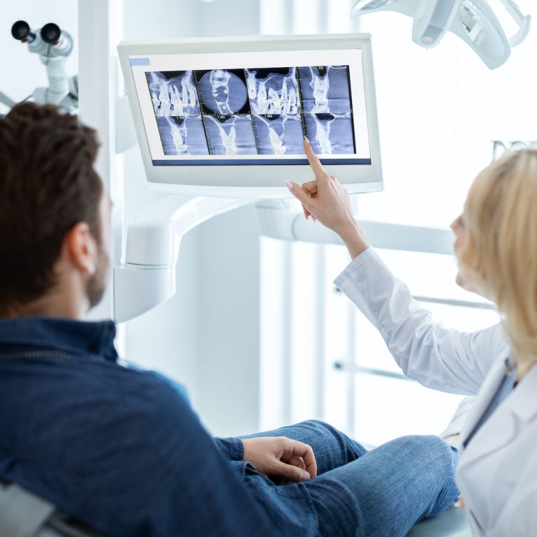 Modern dental clinic concept. Blonde woman dentist having conversation with patient in dental chair, poitning at LED display, showing results of treatment or explaining procedure