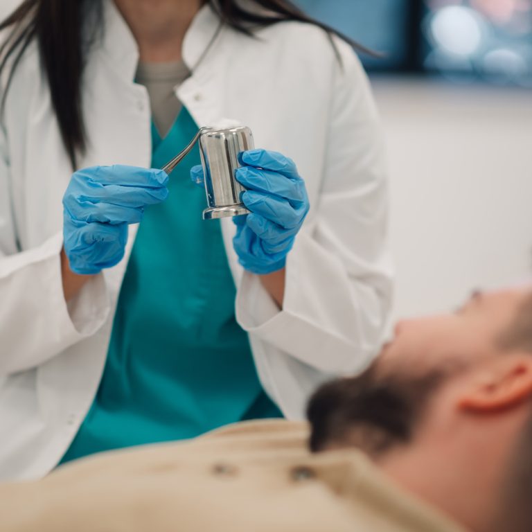 Doctor wearing blue gloves is holding tweezers and a metal container above a patient lying on a medical bed during an intricate procedure in a healthcare setting