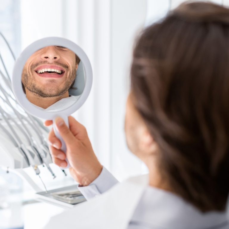Satisfied man looking at mirror and checking his teeth in dental clinic, beautiful smile concept, cropped